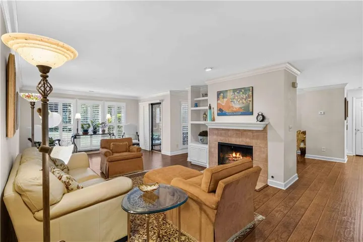 Wood-finish flooring, tiled fireplace with mantel, built-in shelving, white plantation shutters, and crown molding