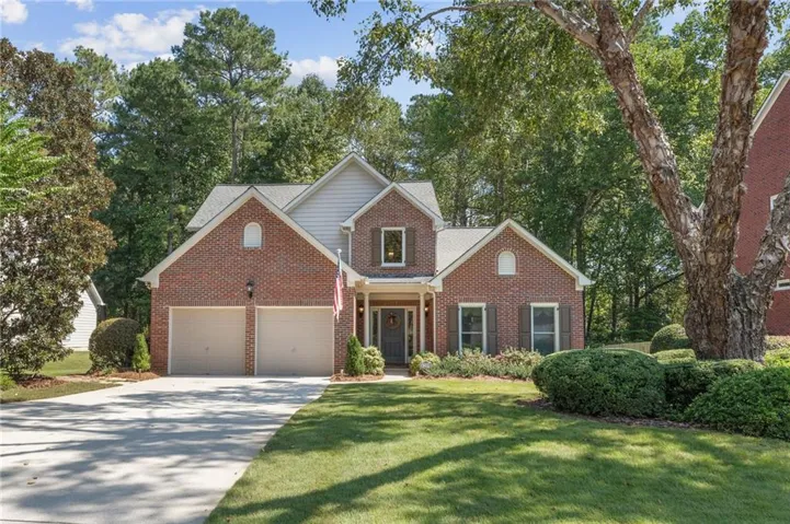 Brick exterior with gabled roof lines, attached two-car garage, manicured lawn, mature trees, and a concrete driveway