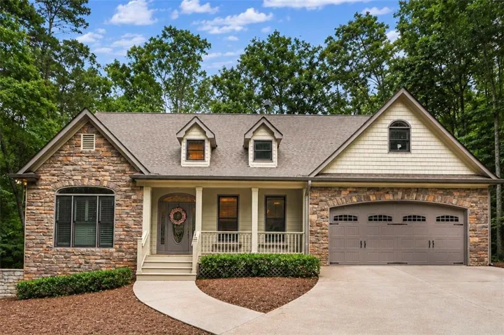 Stone and shake siding facade featuring a covered front porch with white railings, a two-car garage with arched detailing, multiple dormer windows, and a concrete driveway