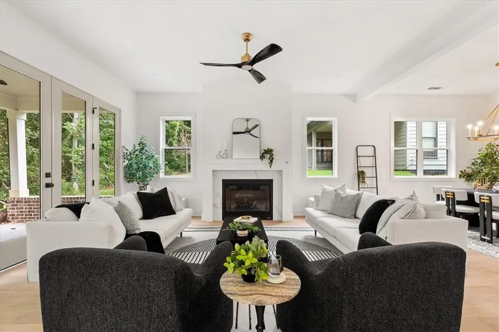 Living area featuring light wood-type flooring, a glass covered fireplace, a chandelier, beam ceiling, and a ceiling fan