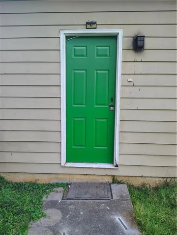 Green panel door with white trim, tan horizontal siding, and a concrete pathway