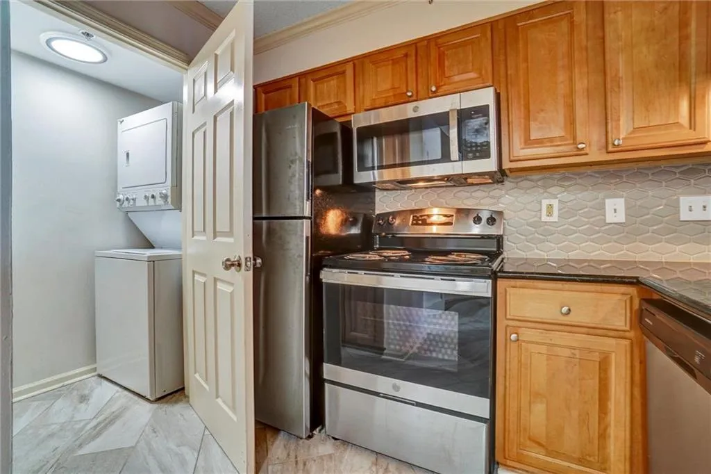 Kitchen with stainless steel appliances, tasteful backsplash, brown cabinetry, dark stone counters, and estacked washer and dryer