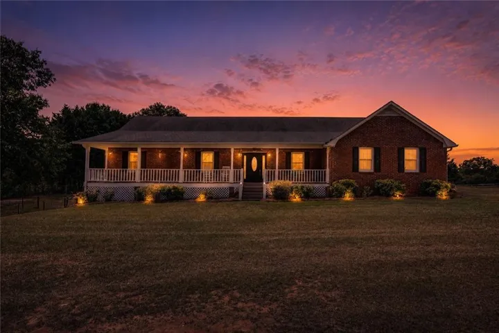 Brick exterior with a covered front porch featuring white railings, landscaping, exterior lighting, and a front entry door with an oval glass insert
