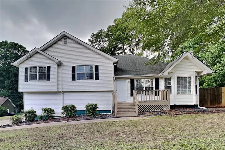 White siding exterior with a dark shingle roof, featuring a front deck with wood railings, a bay window, and dark window shutters