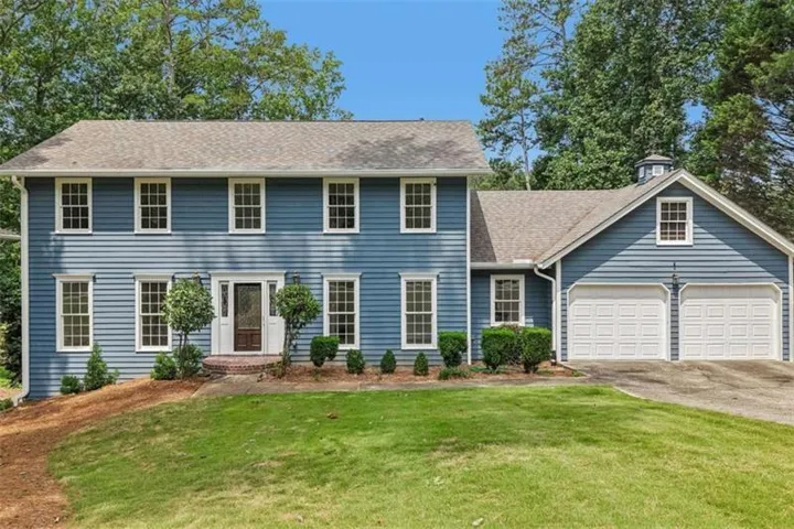 Blue clapboard siding with white trim, two-car garage, brick paver entry, cupola detail, and mature landscaping
