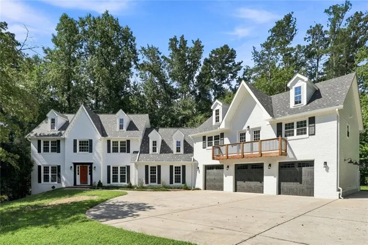 White brick exterior with black shutters, multiple dormer windows, a prominent front entry with an orange door, a second-story balcony, and multiple garage doors