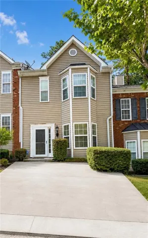 Townhome exterior featuring tan siding with brick accents, bay windows, a white entry door, and a concrete driveway