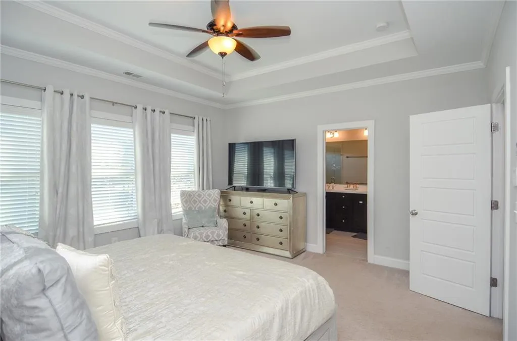 Bedroom with a tray ceiling, light colored carpet, a ceiling fan, ornamental molding, and ensuite bathroom