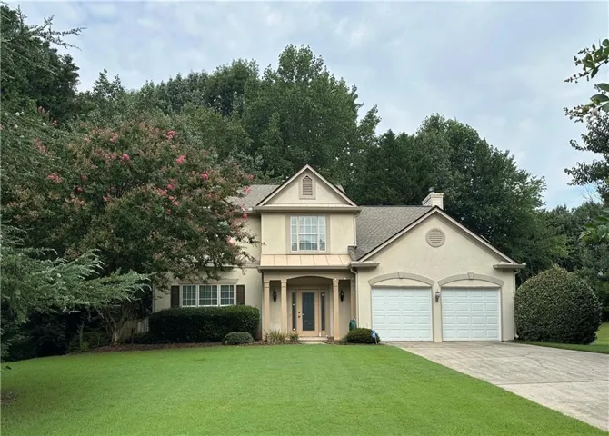 Stucco exterior home featuring a two-car garage, mature landscaping, a symmetrical front façade, a gabled roofline, and a covered front entry