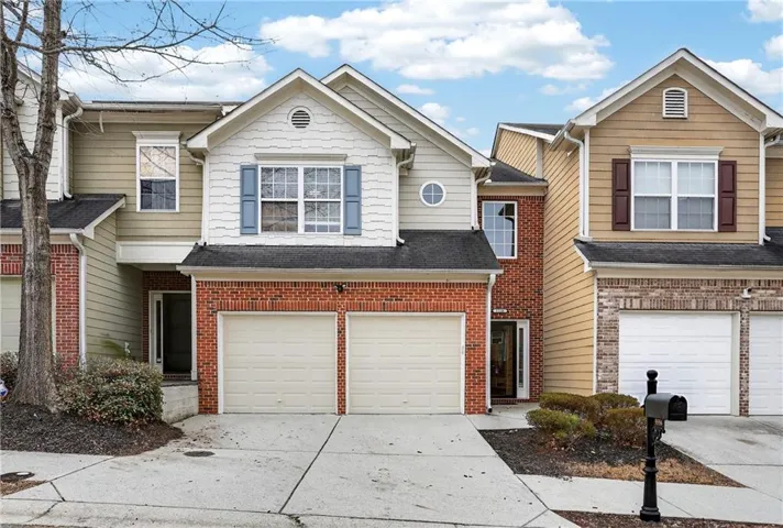 Charming two-story townhome with brick and siding exterior featuring a two-car garage, gabled roofline with circular window detail, and manicured front landscaping with mature trees