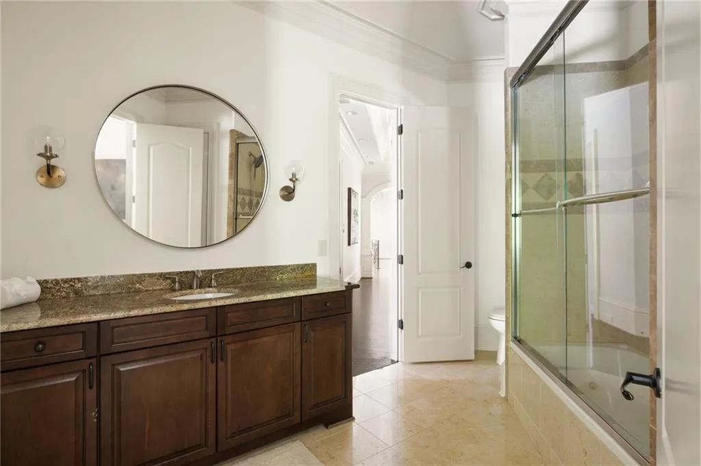 Spacious bathroom featuring an extended vanity with a stone countertop, dark wood-finish cabinetry, a large circular mirror, and a combination tub and shower with glass enclosure