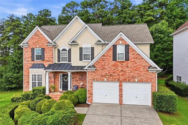 Traditional-style home featuring a brick and stucco facade, a covered entry porch, a two-car garage, mature landscaping, and a shingled roof