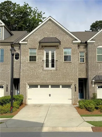 Brick facade with a gabled roofline, featuring a white garage door, concrete driveway, and a balcony with French doors