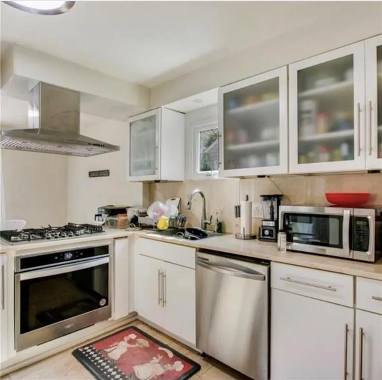 Kitchen featuring stainless steel appliances, wall chimney exhaust hood, light countertops, white cabinetry, and decorative backsplash