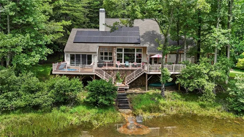 Waterfront cottage nestled among mature trees, featuring a multi-level wood deck with railings, expansive glass-panel doors and windows for natural light, composite or wood-finish exterior siding, and direct water access to a private pond or creek