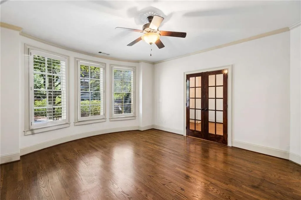 Curved wall with three large windows featuring white plantation shutters, rich wood-finish flooring, dark wood French doors, and a ceiling fan with integrated lighting