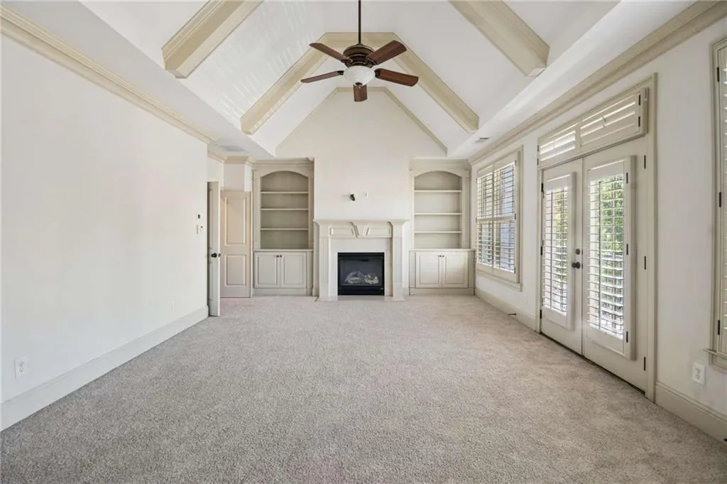 Vaulted ceiling with exposed beams, built-in shelving, fireplace with mantel, double doors with transom, and plantation shutters