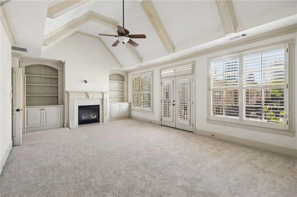 Vaulted ceiling with exposed beams and ceiling fan, built-in shelving with lower cabinetry, central fireplace, and multiple windows with plantation shutters