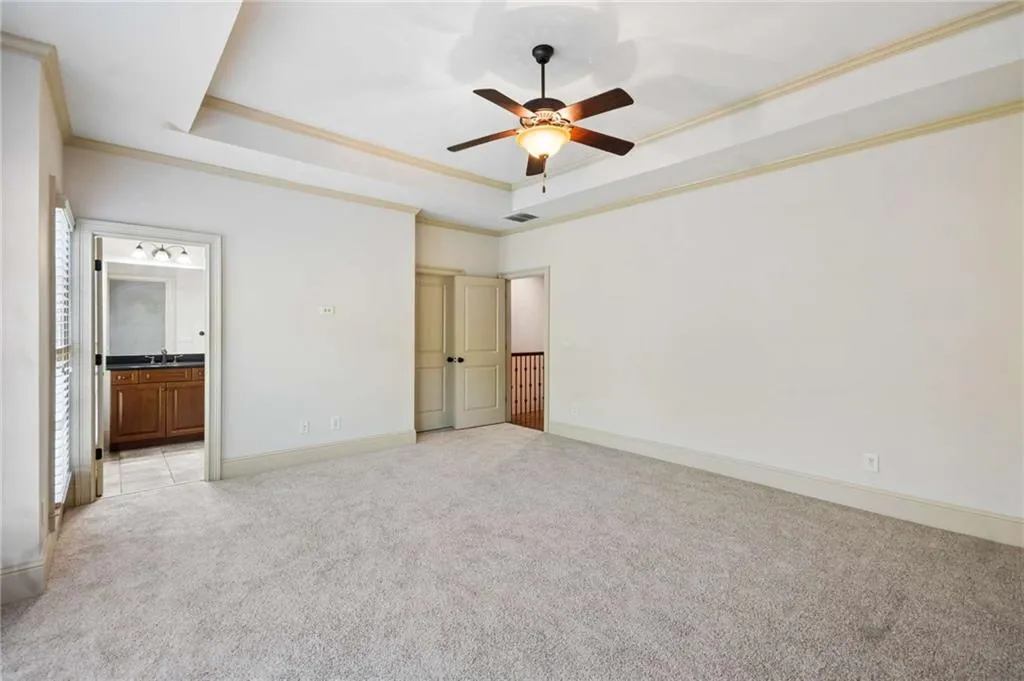 Recessed ceiling with a ceiling fan, light-colored carpet, and an en-suite bathroom with a wood-finish vanity and dark countertop