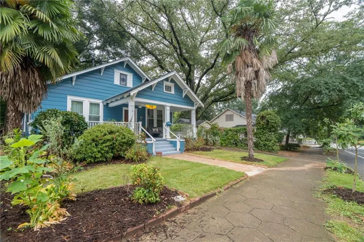 Blue siding with white trim, covered front porch, front yard landscaping, mature trees, and a paved walkway