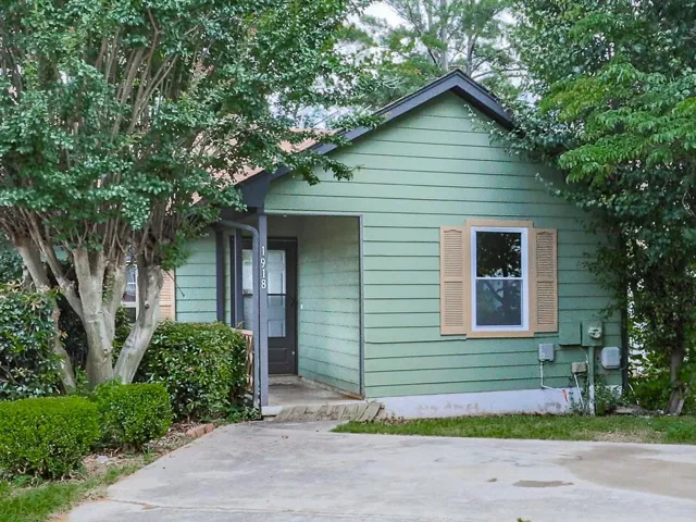 Exterior featuring sage green horizontal siding, a dark gray front door, a single-pane window with tan shutters, and established perimeter shrubbery