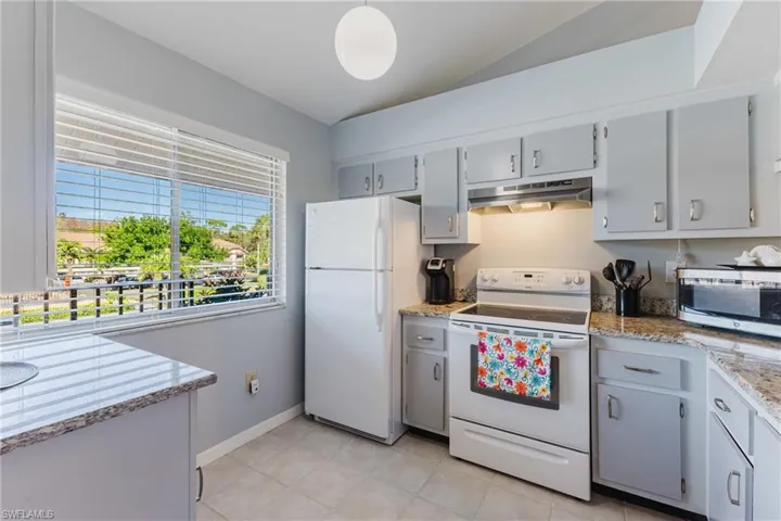 Kitchen with white appliances, light stone countertops, under cabinet range hood, lofted ceiling, and light tile patterned floors