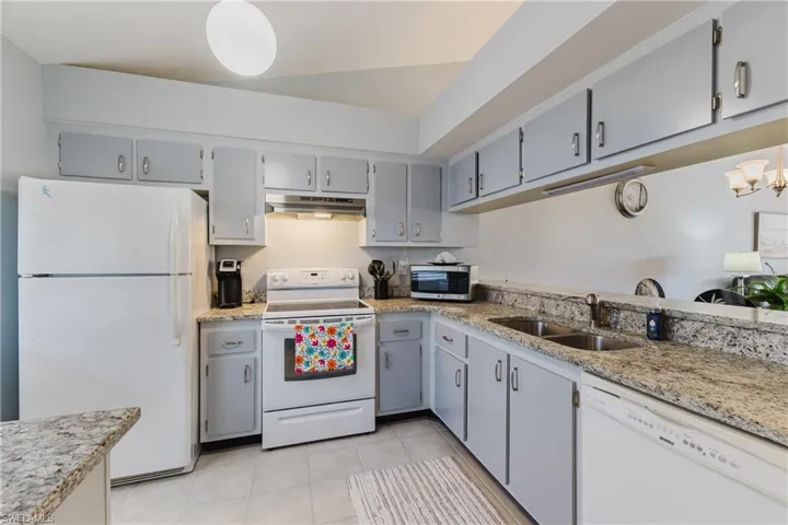 Kitchen with gray cabinetry, white appliances, light stone counters, under cabinet range hood, and a chandelier