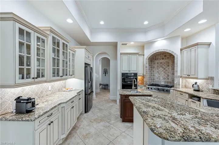Kitchen featuring fridge with ice dispenser, arched walkways, an island with sink, ornamental molding, and decorative backsplash