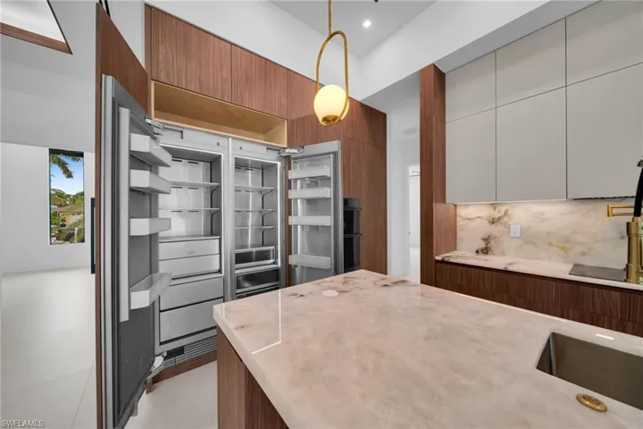 Gourmet kitchen featuring a built-in stainless steel refrigerator, expansive island with an undermount sink, stone countertops and backsplash, custom wood cabinetry, and an architectural pendant light fixture