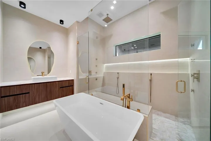 Contemporary bathroom featuring a freestanding soaking tub with brass fixtures, a glass-enclosed shower with multiple showerheads, a floating vanity with a vessel sink, and an organically shaped backlit mirror