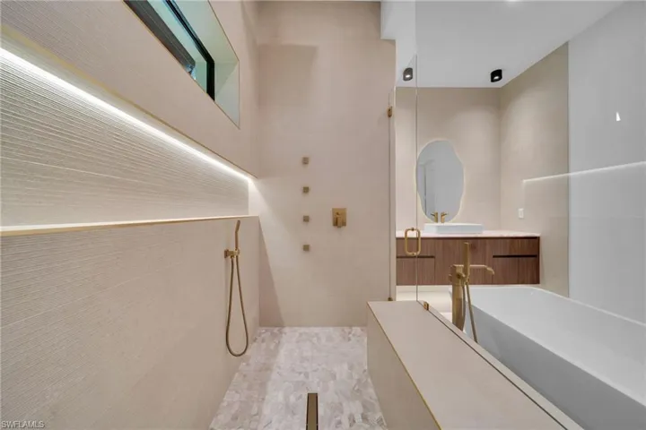 Contemporary bathroom featuring a large walk-in shower with integrated linear lighting, a freestanding soaking tub with floor-mounted faucet, a floating wood-finish vanity with countertop basin, and a uniquely shaped illuminated mirror