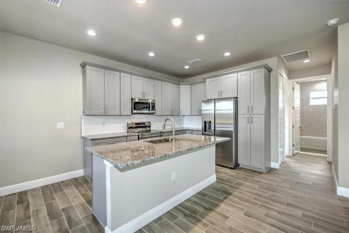 Kitchen with wood tiled floors, stainless steel appliances, gray cabinets, light stone countertops, and an island with sink