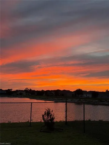 Waterfront property featuring a chain-link fence, mature shrubbery, a grass lawn, and a vibrant sky
