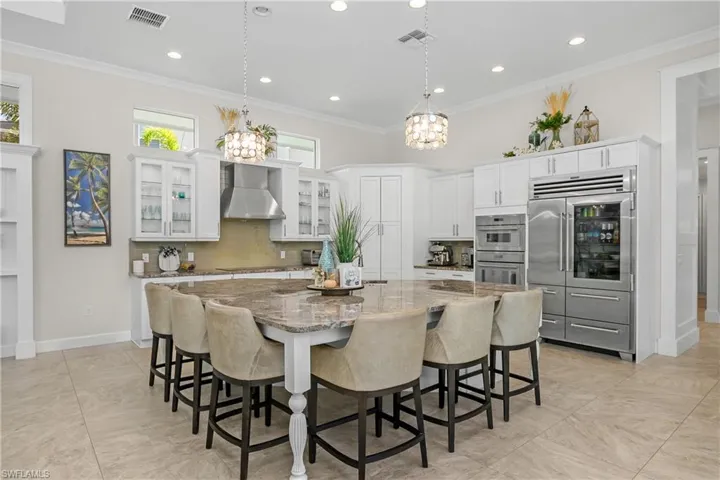 Large Island Kitchen featuring wall chimney exhaust hood, an island with sink, stainless steel appliances, and white cabinetry, pendant lighting, transom windows.
