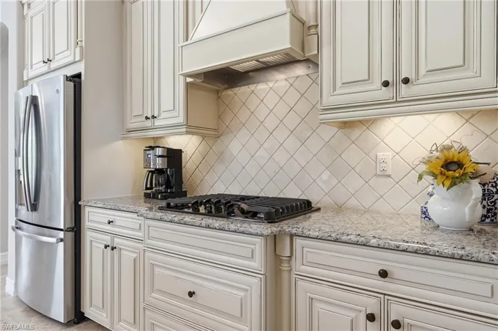 Kitchen featuring cream-toned cabinetry with dark hardware, a stainless steel refrigerator, a built-in gas cooktop, light-toned patterned countertops, and a diamond-set tile backsplash