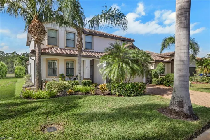 Two-story exterior featuring a light-toned stucco facade, a multi-gabled tile roof, a covered entryway, and a paved driveway, complemented by mature palm trees and manicured landscape shrubbery