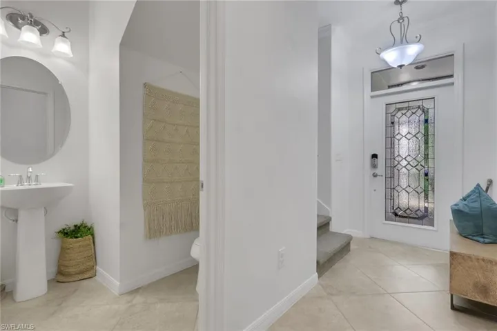 Entryway featuring a front door with a decorative glass insert and transom window, neutral tile flooring, pedestal sink with wall-mounted lighting, and a pendant light fixture