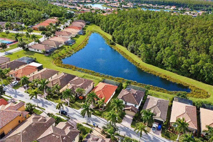 Aerial perspective of the residential community featuring a large retention pond, mature perimeter woodland, paved streets, and residential homes with tile roofing
