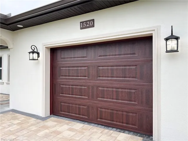 Wood-finish paneled garage door with decorative sconce lighting, bordered by a white stucco exterior and paver driveway