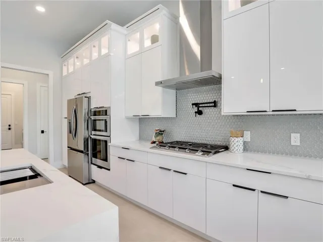 Kitchen featuring tasteful backsplash, appliances with stainless steel finishes, light stone countertops, wall chimney range hood, and white cabinetry