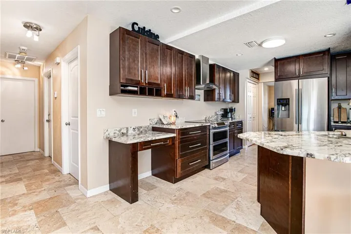 Kitchen with dark brown cabinetry, appliances with stainless steel finishes, light stone countertops, a textured ceiling, and wall chimney exhaust hood
