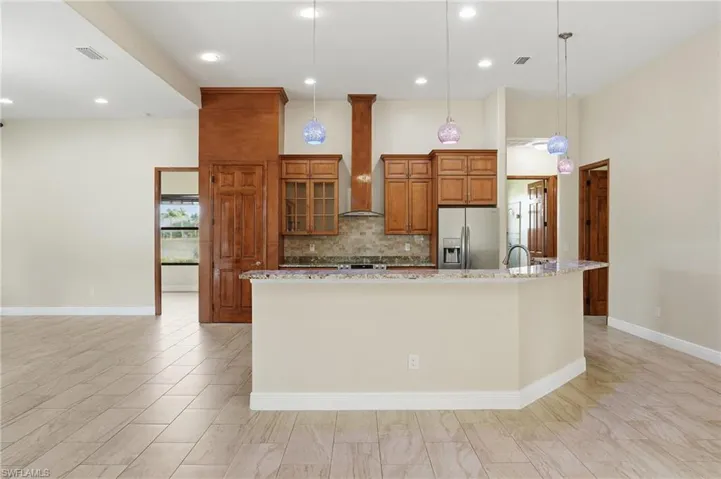 Kitchen with brown cabinets, light stone counters, stainless steel fridge, pendant lighting, and wall chimney range hood