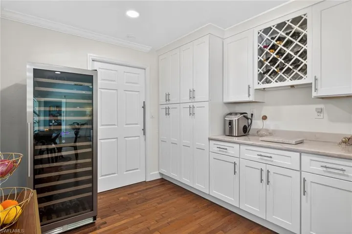 Indoor dry bar with wine cooler, white cabinetry, dark wood finished floors, ornamental molding, and light stone countertops