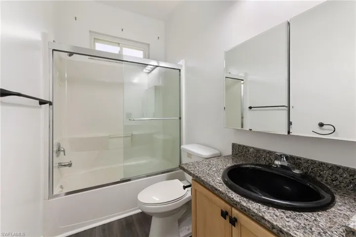 Full bathroom featuring a granite-style countertop with an oval black sink, a light wood-finish vanity, and a shower-tub combination with sliding glass doors