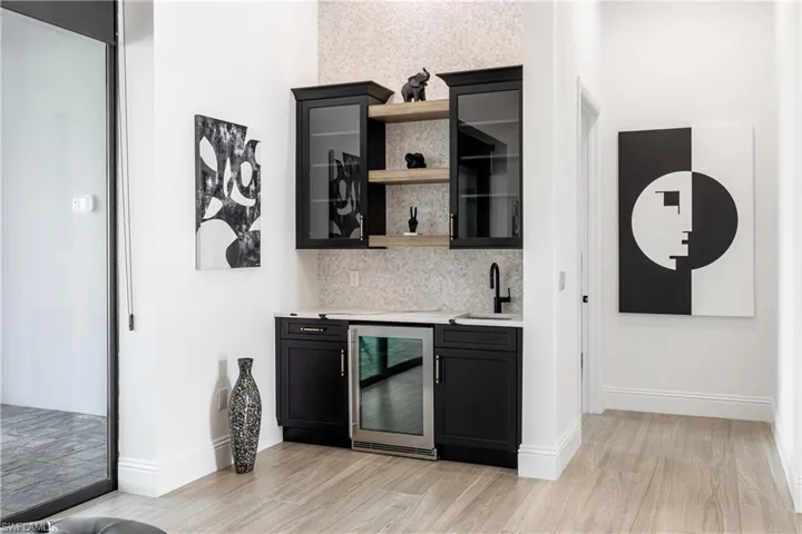 Indoor wet bar with dark cabinets, open shelves, wine cooler, glass fronted cabinets, and backsplash