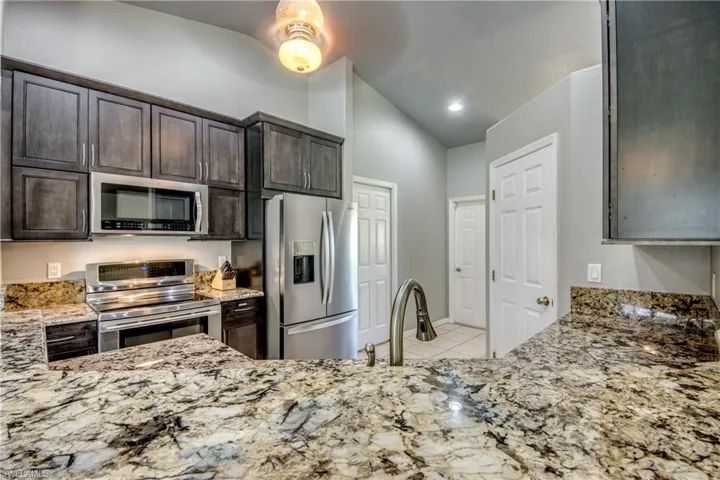 Kitchen featuring appliances with stainless steel finishes, vaulted ceiling, light stone countertops, dark brown cabinets, and light tile patterned floors