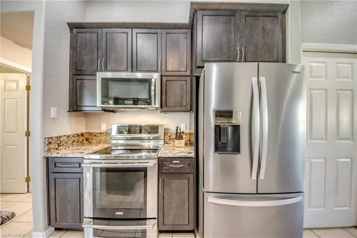Kitchen with stainless steel appliances, light tile patterned flooring, dark brown cabinets, light stone countertops, and a textured wall