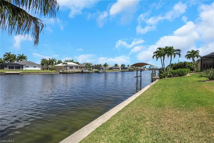 Waterfront property featuring a concrete seawall, lush green lawn, mature palm trees, and a private dock with a covered boat lift