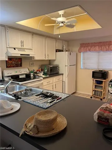 Kitchen with white appliances, light tile patterned floors, under cabinet range hood, dark countertops, and ceiling fan