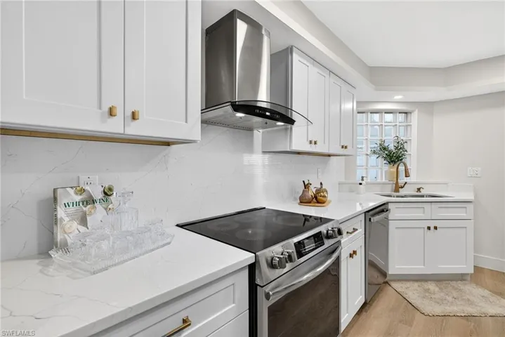 Kitchen featuring stainless steel appliances, wall chimney range hood, light stone countertops, and white cabinetry